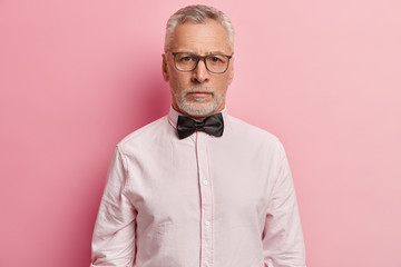 Portrait of self assured bearded man with grey hair, wrinkled face, dressed in shirt with bowtie, thinks about retirement, poses over rosy background, wants to share his wisdom with grandchildren