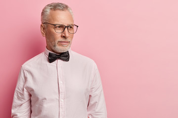 Studio shot of handsome male pensioner being deep in thoughts, focused away, has clever look, thinks how develop his business, wears glasses and formal outfit, isolated over pink background.