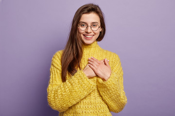Smiling joyous woman with straight dark hair, touches by impressive words, holds palms on heart, expresses sympathy and good feeling, wears knitted yellow jumper, isolated on purple studio wall