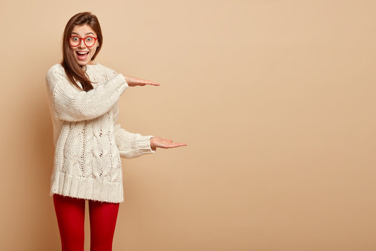 Half length shot of overjoyed pleased woman smiles happily, shapes big object with both hands, wears sweater and red leggings, demonstrates large invisible item, isolated over beige studio wall