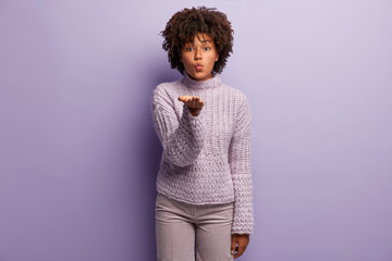 Body language concept. Pretty dark skinned woman stretches hand over chest, blows air kiss at camera, expresses love to people, wears knitted jumper, isolated over purple background. Mwah to you