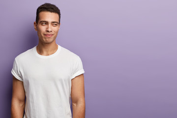 Horizontal shot of handsome European man has muscular body, wears white t shirt with empty space, keeps gaze away, isolated over purple background with empty space for your promotional text.