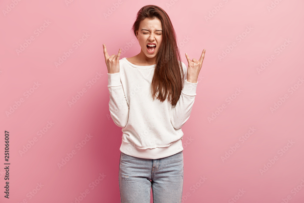 Wall mural Positive emotional woman makes rock n roll gesture, exclaims loudly while listens loud music, dressed in casual jumper and jeans, isolated over pink background. I am fun of rock. Horizontal shot