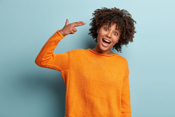 Waist up shot of good looking funny Afro American young woman shoots in temple, tilts head, pretends killing herself, dressed in orange jumper, isolated over blue background. Suicide gesture