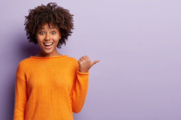 Glad girl with crisp black hair, dark skin, toothy smile, points with thumb aside, demonstrates something, shows copy space for your usage, wears orange jumper, isolated over purple background