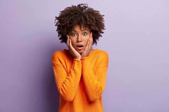 Terrific dark skinned woman touches cheeks, opens eyes with surprirsement, keeps jaw dropped, has Afro haircut, wears orange jumper, models against purple background, hears something astonishing