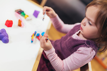 Kid girl is playing with plasticine while sitting at table in nursery room, selective focus.