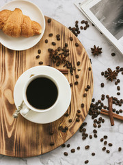Cup of black coffee with tasty croissant on a tray on a marble table with spices and fashion magazine. Author processing, film effect.