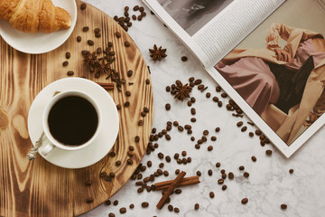 Cup of black coffee with tasty croissant on a tray on a marble table with spices and fashion magazine. Author processing, film effect.