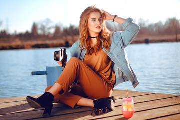 Beautiful girl sitting on the pier near the river. Hipster model in choker with blond cerly hair. The girl at hot sunny day taking pictures old camera, Summer vibes. Woman photographer hobby.