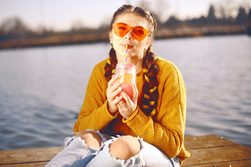 The girl at hot sunny day drink lemonad. Summer vibes. Girl with fashion yellow sunglasses and dressed in yellow. Beautiful girl sitting on the pier near the river. Beauty model with brunette hair.