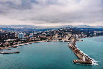 Fototapeta na wymiar Aerial view of Yalta embankment from drone, old Lighthouse on pier, sea coast landscape and city buildings on mountains, beautiful winter panorama of European resort, Crimea