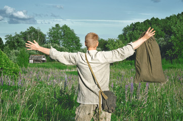man in an old military uniform with a large bag standing in the field with his arms outstretched