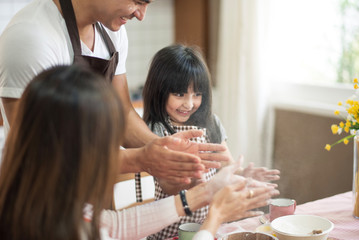 The family cooks. Daughter, Mother and Father prepare dough powder. All have aprons. Live at home in the kitchen. They enjoy and smile.