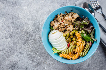 Healthy lunch bowl. Chicken, pasta fusilli, mix greens, green peas and sunflower seeds in blue plate on concrete gray background table. Delicious balanced food concept. Top view, copy space
