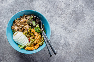 Healthy lunch bowl. Chicken, pasta fusilli, mix greens, green peas and sunflower seeds in blue plate on concrete gray background table. Delicious balanced food concept. Top view, copy space