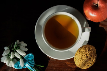 White cup with tea on a saucer with oatmeal cookies and red apple and a small bouquet of snowdrops tied with a blue ribbon