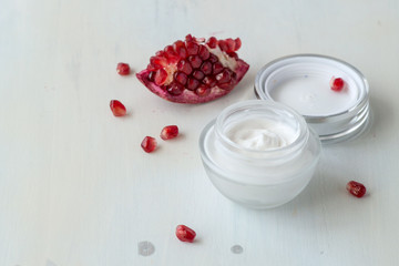 A small jar with a cream for face skin care and a piece of ripe red pomegranate on a light wooden background.Selective focus.