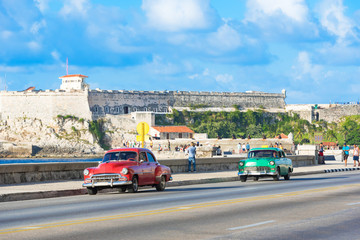Amerikanischer roter und grüner Oldtimer unterwegs auf dem berühmten Malecon und im Hintergrund die Festung Castillo de los Tres Reyes del Morro in Havanna Kuba - Serie Kuba Reportage