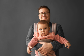 Fathre and his kid in baby carrier at the grey background wall, baby wearing concept