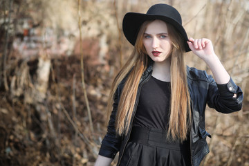 A girl walks down the city street in a leather waistcoat with a phone. Young beautiful girl in a hat and with a dark make-up outside. Girl in the Gothic style on street.