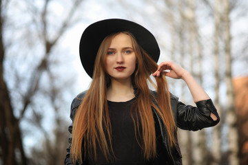 A girl walks down the city street in a leather waistcoat with a phone. Young beautiful girl in a hat and with a dark make-up outside. Girl in the Gothic style on street.