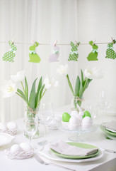Decor and table setting of the Easter table with white tulips and dishes of green and white color. Easter decor in the form of Easter bunnies  green color with white polka dots.
