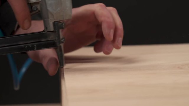 A side view of a man's hand holding pneumatic staple gun and stapling a plywood board to wooden drawer