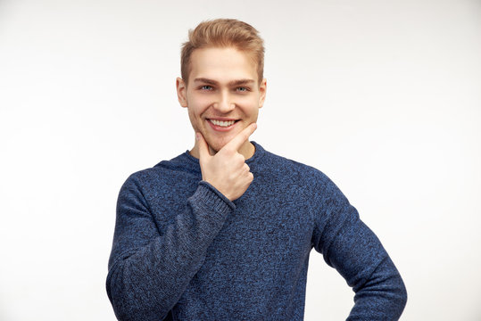 Horizontal  fhoto of confident bearded male keeps finger on chin, has attentive look, dressed in blue jumper. Self assured businessman looks seriously, poses against white background