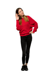 Fototapeta premium A full-length shot of a Young woman with red sweater listening to something by putting hand on the ear over isolated white background