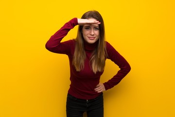 Woman with turtleneck over yellow wall looking far away with hand to look something
