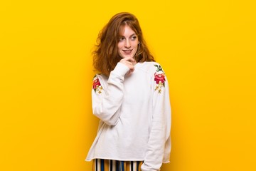 Young redhead woman over yellow wall looking to the side with the hand on the chin