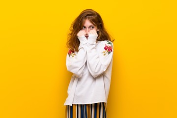 Young redhead woman over yellow wall is a little bit nervous and scared putting hands to mouth