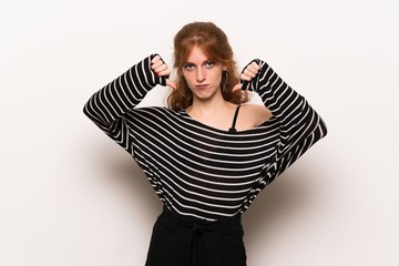 Young redhead woman over white wall showing thumb down with both hands
