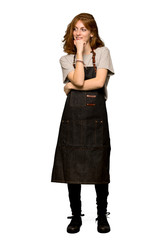 A full-length shot of a Young redhead woman with apron looking to the side with the hand on the chin over isolated white background