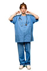 A full-length shot of a Young redhead nurse covering both ears with hands over isolated white background