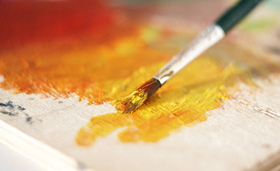 The wooden palette is painted with yellow and red oil paint. The brush blends the paint in orange color.