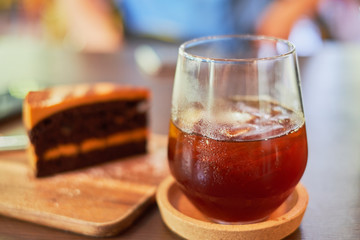 Ice black coffee in glass on foreground and soft blur for chocolate caramel cheese cake serve on wood plate