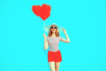 Happy smiling young woman with red heart shaped air balloons in summer straw hat and shorts on colorful blue background