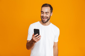Photo of attractive man 30s in casual wear smiling and holding smartphone, isolated over yellow...