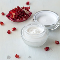 A small jar with a cream for face skin care and a piece of ripe red pomegranate on a light wooden background.Selective focus.