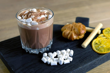 Cocoa with marshmallow in rock glass on a wood black tray with cake and lemon. Close-up side view.