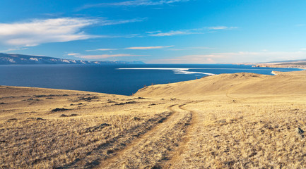 Fototapeta na wymiar Siberian Lake Baikal. Typical steppe landscape of Olkhon Island in spring at sunset. Dirt road on a hillside. Khuzhir village is visible in the distance
