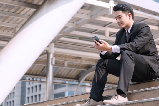 Handsome young businessman wearing black suit sitting on stair using smartphone in modern city background. Professional young man holding cellphone in his hand working online on outdoor.