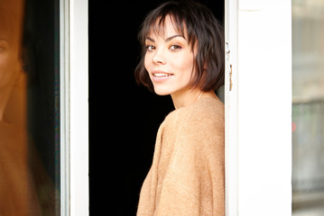 Attractive young woman standing at window while looking at camera and smiling