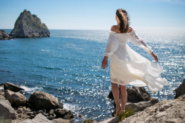 Fototapeta na wymiar beautiful dark-haired young woman in white light dress with long legs stands or dancing at the edge of a rock above the sea, with blue sky and sea background 