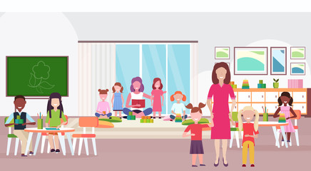 women teachers teaching mix race boys and girls preschool modern kindergarten children classroom with chalkboard desks chairs playground kid room interior full length flat horizontal