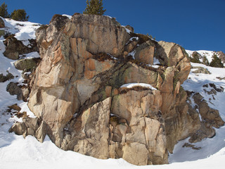 A cliff on a snowy slope against the blue sky with a lone tree on top.
