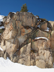 A cliff on a snowy slope against the blue sky with a lone tree on top.