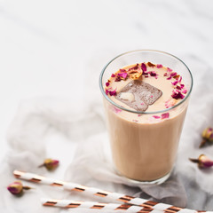 Iced coffee with rose and cardamom in a tall glass surrounded by dried roses and straws on white and gray silk background. Copy space for text. Square crop.